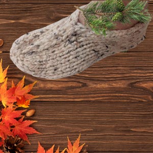 May include: A chunky, hand-knitted slipper in a speckled cream yarn. The slipper is filled with a sprig of green pine needles and small pine cones. Autumn leaves in red and orange and a pine cone are arranged on a wooden surface.