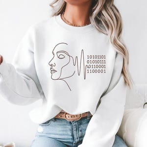 Może przedstawiać: Biała bluza z minimalistycznym rysunkiem linii profilu kobiety i sekwencją kodu binarnego w kolorze ciemnobrązowym. Bluza ma długie rękawy i okrągły dekolt.