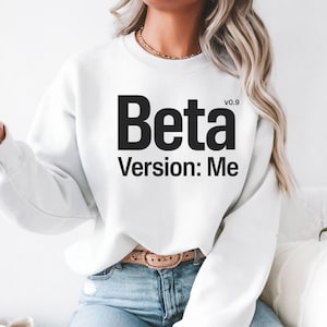 以下が含まれることがあります： 前面に「Beta Version: Me」と黒字でプリントされた白いスウェットシャツ。「Beta」の文字は大きく太字で、「v0.9」が上にあります。「Version: Me」のフレーズは下にあります。