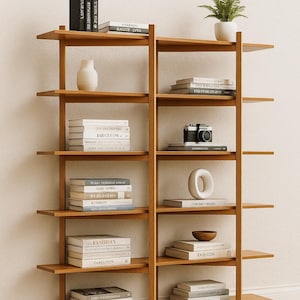 Puede incluir: Una estantería de madera con múltiples estantes, que muestra libros, una cámara, una pequeña planta y objetos decorativos. Los estantes están llenos de libros con títulos como "FASHION" y "The Photography".