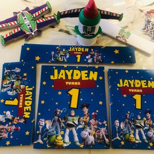 Op de afbeelding: Een set Toy Story-thema feestdecoraties met een blauwe achtergrond met sterren, de personages Woody, Buzz Lightyear, Jessie en anderen. De decoraties omvatten een banner met de tekst "Jayden wordt 1" en verschillende vierkante kaarten met hetzelfde ontwerp en dezelfde tekst.