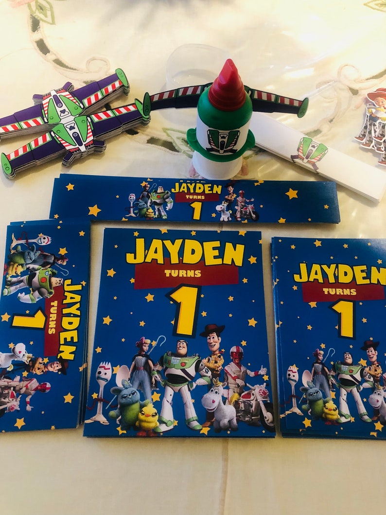 Op de afbeelding: Een set Toy Story-thema feestdecoraties met een blauwe achtergrond met sterren, de personages Woody, Buzz Lightyear, Jessie en anderen. De decoraties omvatten een banner met de tekst "Jayden wordt 1" en verschillende vierkante kaarten met hetzelfde ontwerp en dezelfde tekst.