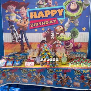 Op de afbeelding: Een verjaardagsfeesttafel opgezet met een Toy Story-thema. De tafel is bedekt met een blauwe tafelkleed met een Toy Story-ontwerp. Er staan snacks, drankjes en cupcakes op de tafel. De achtergrond is een Toy Story-banner met de woorden "Happy Birthday".