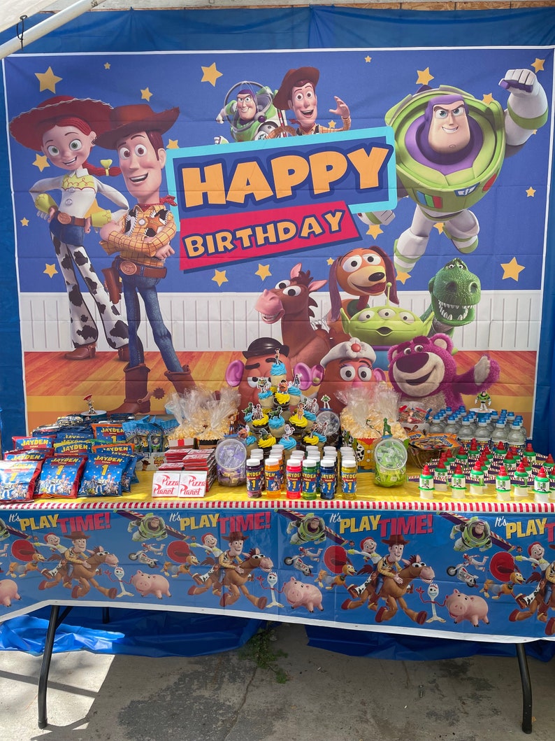 Op de afbeelding: Een verjaardagsfeesttafel opgezet met een Toy Story-thema. De tafel is bedekt met een blauwe tafelkleed met een Toy Story-ontwerp. Er staan snacks, drankjes en cupcakes op de tafel. De achtergrond is een Toy Story-banner met de woorden "Happy Birthday".