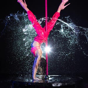 May include: A bright pink dance pole set against a black background with water splashing around the base. The attire includes neon pink joggers, a matching pink sports bra, and light pink platform high heels with ankle straps and fishnet stockings.