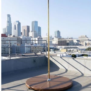 May include: A vertical metallic pole with a polished finish, mounted securely into the center of a circular, brown, padded base. The structure stands upright on a flat surface, featuring a silver-toned connector at the base of the pole.