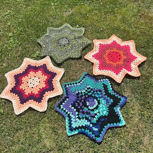 May include: Four star-shaped crochet mats featuring intricate yarn patterns. One mat displays shades of green, another features pink and orange, a third shows purple and peach, and the fourth combines black, blue, and teal yarn colors.
