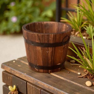 Könnte beinhalten: Ein kleiner, runder Holzblumentopf mit dunkelbraunem Holz und zwei schwarzen Metallbändern. Der Topf steht auf einer Holzoberfläche, im Hintergrund grüne Pflanzen. Der Topf ist für kleine Pflanzen oder Blumen gedacht.