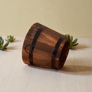 Puede incluir: Un cubo de madera rústico con aros de metal oscuro y un asa. El cubo está hecho de madera teñida y tiene forma cónica. El cubo está vacío y descansa sobre una superficie de madera de color claro.