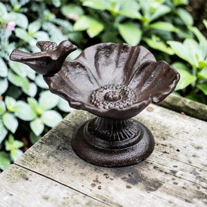 Puede incluir: Un bebedero para pájaros de hierro fundido con acabado marrón oscuro. El cuenco tiene forma de flor, con un pequeño pájaro en el lateral. La base es redonda y decorativa. El bebedero está sobre una superficie de madera, rodeado de vegetación.