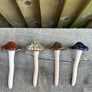 May include: Four ceramic mushroom garden stakes with varying cap colors: red, tan, brown, and blue. Each mushroom has a white or pink stem. The stakes are arranged on a weathered wooden surface.