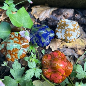 May include: Four colorful, organic-shaped ceramic stones. One is orange with turquoise spots, another is blue with green spots, a third is brown with white spots, and the last is red with orange spots. The stones are surrounded by green leaves and brown foliage.