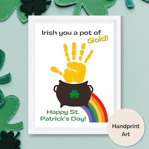 May include: A white card with a yellow handprint inside a black pot with a green shamrock. The text reads "Irish you a pot of Gold!" and "Happy St. Patrick's Day!" with a rainbow underneath the pot. The card has a white frame and a white circle with the text "Handprint Art" in the bottom right corner.