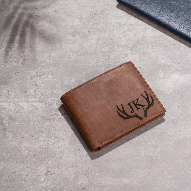 May include: A brown leather wallet with a bi-fold design. The wallet features a deer antler graphic and the initials "JK" engraved on the front. The wallet is shown on a gray surface.