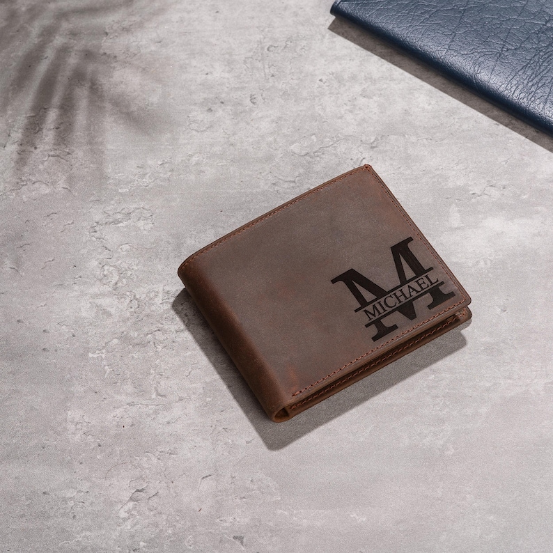 May include: A brown leather wallet with the name "MICHAEL" and the letter "M" engraved on the front. The wallet is rectangular and appears to be folded. The background is a light gray surface.