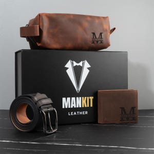 May include: A brown leather toiletry bag, wallet, and belt set with a black box. The box has a white tuxedo graphic and the words "MANKIT LEATHER". The wallet and toiletry bag are personalized with the letter "M" and the name "MICHAEL".
