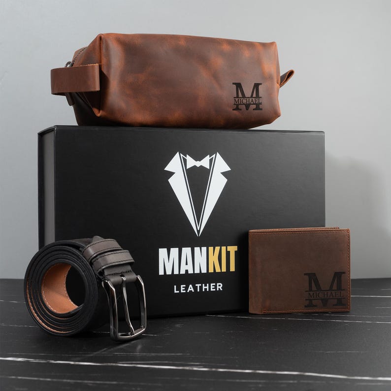 May include: A brown leather toiletry bag, wallet, and belt set with a black box. The box has a white tuxedo graphic and the words "MANKIT LEATHER". The wallet and toiletry bag are personalized with the letter "M" and the name "MICHAEL".