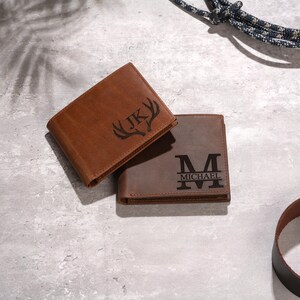 May include: Two brown leather wallets. One is a lighter shade of brown with the initials "JK" and a deer antler design. The other is a darker brown with the letter "M" and the name "MICHAEL".