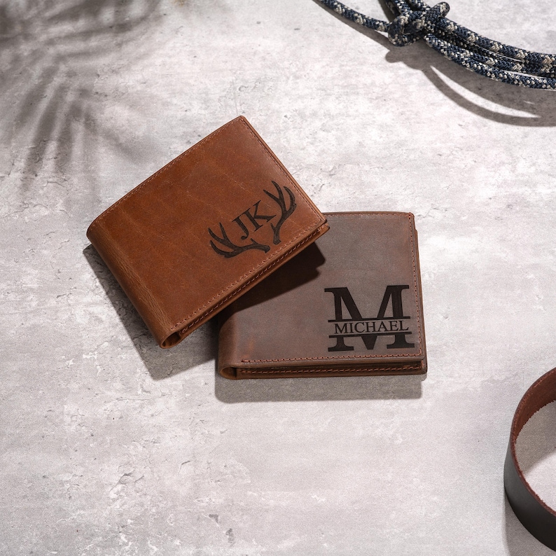 May include: Two brown leather wallets. One is a lighter shade of brown with the initials "JK" and a deer antler design. The other is a darker brown with the letter "M" and the name "MICHAEL".