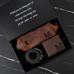 May include: A gift box containing a brown leather wallet, belt, and travel bag, all with the initial "M" and the name "MICHAEL" engraved. The box also includes a message: "Thank you for being part of what we love to do."