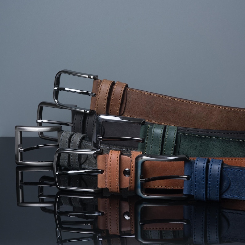 May include: A collection of leather belts in various colors, including brown, black, green, tan, and blue. Each belt features a silver-colored metal buckle. The belts are arranged in a stacked display, showcasing their different hues and textures.