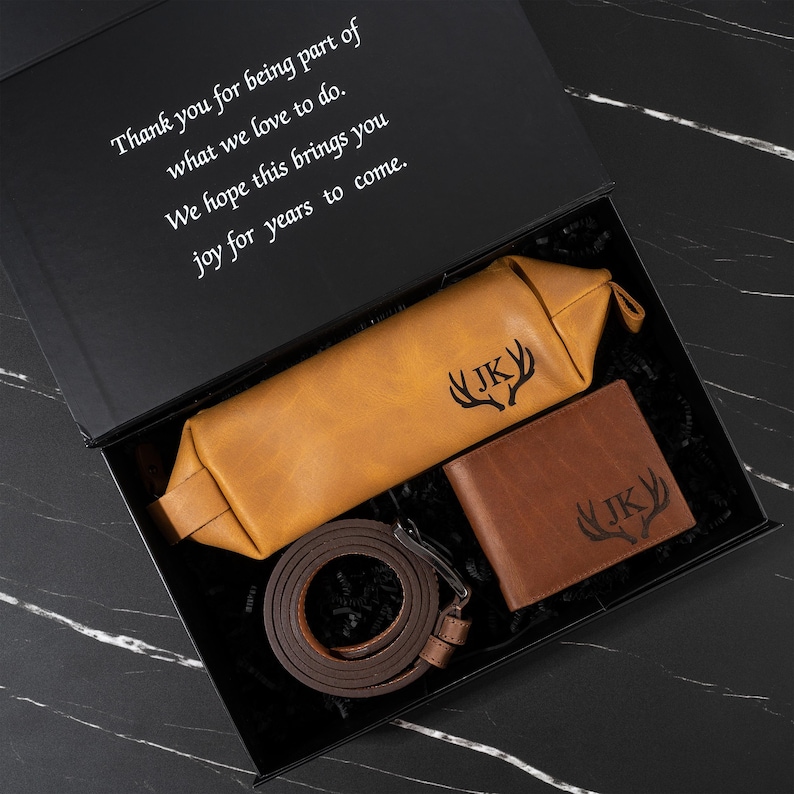 May include: A gift set in a black box, featuring a tan leather toiletry bag, a brown leather wallet, and a brown leather belt. The toiletry bag and wallet are embossed with "JK" and a deer antler design. The box lid has a printed message.