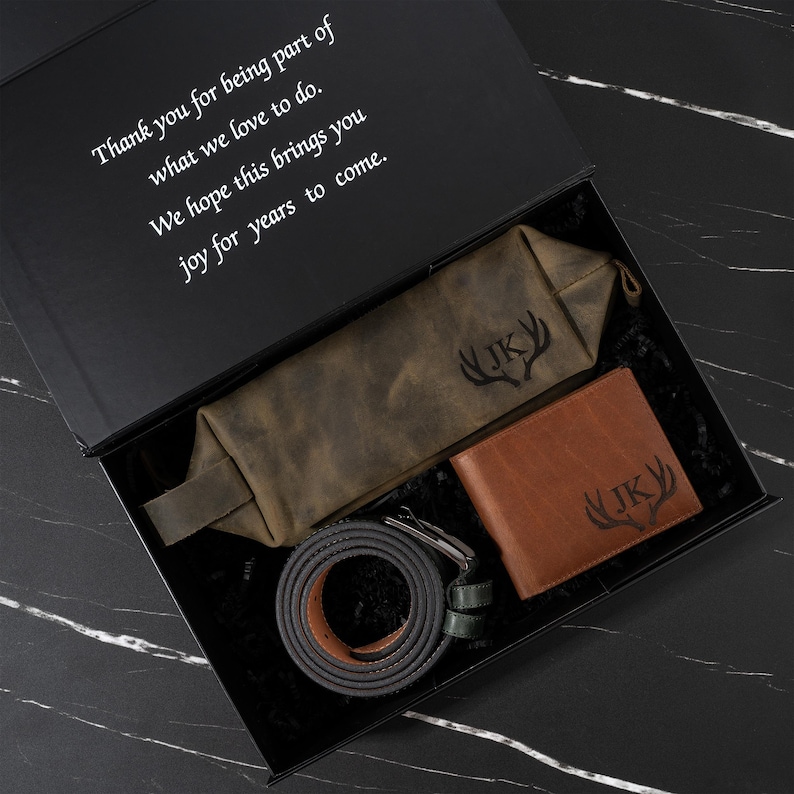 May include: A gift set in a black box, including a brown leather wallet, a green pouch, and a gray belt. The wallet and pouch are embossed with "JK" and a deer antler design. The box lid has white text that reads "Thank you for being part of what we love to do. We hope this brings you joy for years to come."
