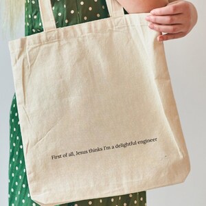 May include: A beige canvas tote bag featuring the black printed text, First of all, Jesus thinks I'm a delightful engineer. The bag has two shoulder straps and is constructed from a plain, light-colored fabric with a simple rectangular shape.
