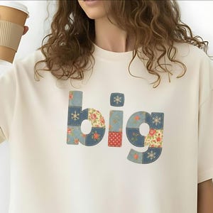 Peut inclure: T-shirt oversize ivoire avec le mot "big" en lettres patchwork. Les lettres sont composées de motifs floraux et flocons de neige en bleu, rouge et jaune. Le texte "Ivory size up for the oversized look" est en bas.