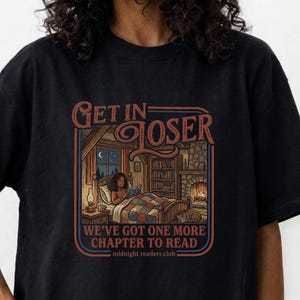 May include: Black t-shirt with a graphic that reads "Get in Loser" above an illustration of a person reading in bed. The text "We've got one more chapter to read" is below the illustration. The shirt is black.