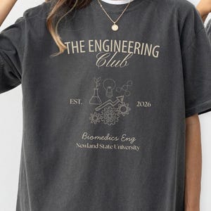 May include: Dark grey t-shirt featuring the text "THE ENGINEERING Club" and a graphic of a lightbulb, gears, and scientific symbols. The shirt also includes the text "EST. 2026" and "Biomedics Eng Newland State University". The shirt is a pepper colour.