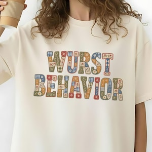 May include: An ivory short-sleeved t-shirt featuring the text WURST BEHAVIOR printed in a multi-colored patchwork pattern. The letters are filled with various floral, gingham, and geometric designs in shades of blue, red, yellow, and green.