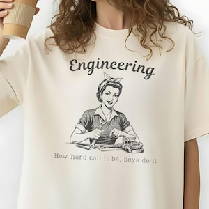 May include: An ivory oversized t-shirt featuring the word "Engineering" and a vintage illustration of a woman working on machinery. The text "How hard can it be, boys do it" is below the illustration. The text "Ivory size up for the oversized look" is at the bottom.