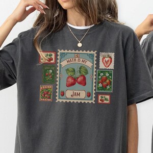 Peut inclure: T-shirt gris foncé avec un motif sur le thème des fraises et le texte "MATH IS MY JAM" dans un design de timbre. Le mot "FRAISE" est également imprimé sur le t-shirt. Fait partie d'une collection de vêtements.