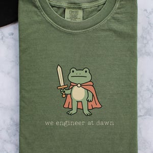 May include: Moss green t-shirt featuring a cartoon frog in a cape, holding a sword. The text "we engineer at dawn" is printed below the frog. The shirt is folded and has a Comfort Colors label.