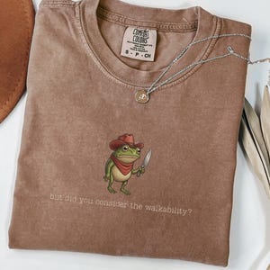 May include: Espresso-colored t-shirt with a graphic of a frog wearing a cowboy hat and holding a knife. The text "but did you consider the walkability?" is printed below the frog. A silver necklace with a pendant is draped across the shirt.