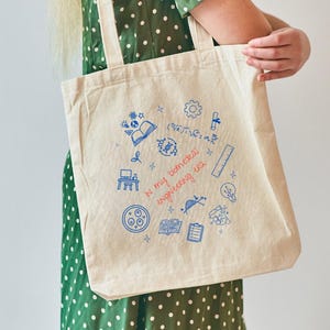 May include: A beige canvas tote bag with blue and red illustrations and text. The bag features small drawings of books, gears, and a pizza. The text on the bag reads "in my dreams, engineering life".