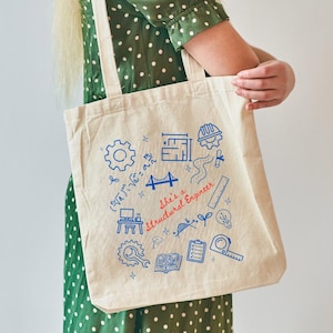 May include: A beige canvas tote bag with blue and red illustrations of engineering tools and the text "She's a Structural Engineer". The bag has two white straps and is held by a person wearing a green dress.