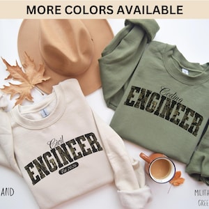 Peut inclure: Deux sweatshirts, l'un sable et l'autre vert militaire, avec les mots "Civil Engineer" et "Coding Engineer" imprimés. Le texte a un motif camouflage. Le sweat-shirt sable porte l'inscription "Est 2026". Un chapeau marron et une feuille d'automne sont également présents.