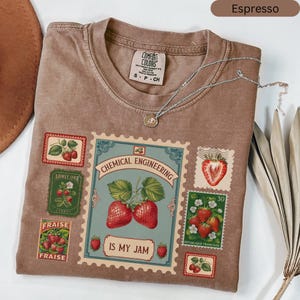 May include: Espresso-coloured t-shirt with strawberry postage stamp graphics. The shirt features the text "Chemical Engineering Is My Jam" and a silver necklace with a pendant.