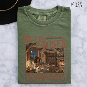 Op de afbeelding: Mosgroen t-shirt met de tekst "GET IN LOSER" boven een illustratie van een persoon die in bed leest. De illustratie bevat een open haard, boekenplanken en een kat. De tekst "WE'VE GOT ONE MORE CHAPTER TO READ" staat eronder.
