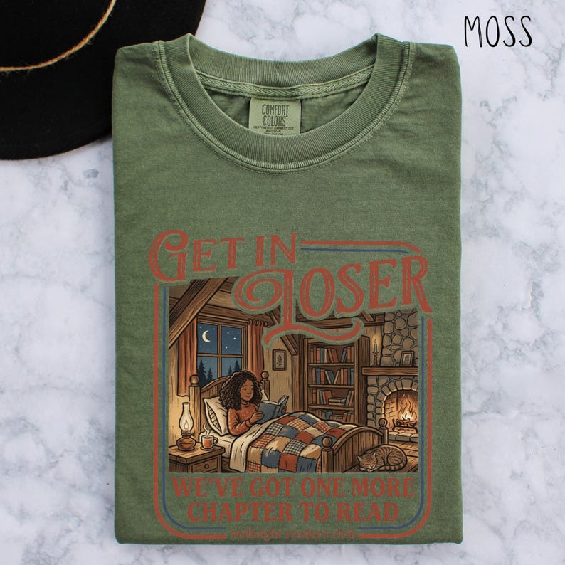 Op de afbeelding: Mosgroen t-shirt met de tekst "GET IN LOSER" boven een illustratie van een persoon die in bed leest. De illustratie bevat een open haard, boekenplanken en een kat. De tekst "WE'VE GOT ONE MORE CHAPTER TO READ" staat eronder.