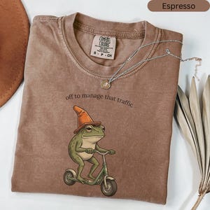 May include: Espresso-colored t-shirt with a frog graphic riding a scooter and wearing a cone hat. The text "off to manage that traffic" is above the frog. A silver necklace with a pendant is draped across the shirt.