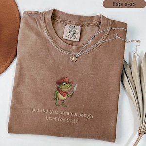 May include: Espresso-coloured t-shirt featuring a cartoon frog in a cowboy hat, holding a knife, with the text "but did you create a design brief for that?". The shirt has a Comfort Colors label and a silver necklace with a pendant.
