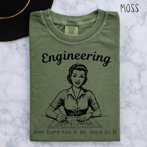 May include: Moss green t-shirt featuring the word "Engineering" and a black and white illustration of a woman working on a blueprint. The text "How hard can it be, boys do it" is below the illustration. The shirt is made of a soft material.