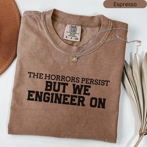 May include: Espresso-coloured t-shirt with the text "THE HORRORS PERSIST BUT WE ENGINEER ON" in black. A silver necklace with a circular pendant rests on the shirt. The shirt has a crew neck and a Comfort Colors tag.