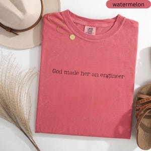 May include: A watermelon-coloured t-shirt with the text "God made her an engineer" in black. A gold necklace with a circular pendant rests on the shirt. A beige hat and sandals are also visible.