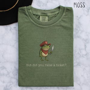 May include: Moss green t-shirt featuring a cartoon frog in a cowboy hat, holding a knife, with the text "but did you raise a ticket?". The shirt has a Comfort Colors label. The shirt is a moss green colour.