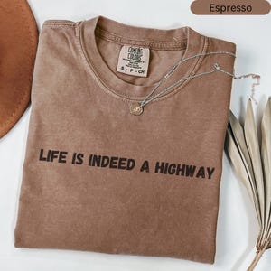 May include: Espresso-coloured t-shirt with the text "LIFE IS INDEED A HIGHWAY" in black. A silver necklace with a pendant rests on the shirt. The shirt has a crew neck and a Comfort Colors tag.