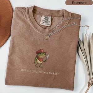 May include: A white ceramic mug with a black handle and the text "But First, Coffee" in a bold, black font. The mug is set against a blurred background. The mug appears to be approximately 10 cm tall.